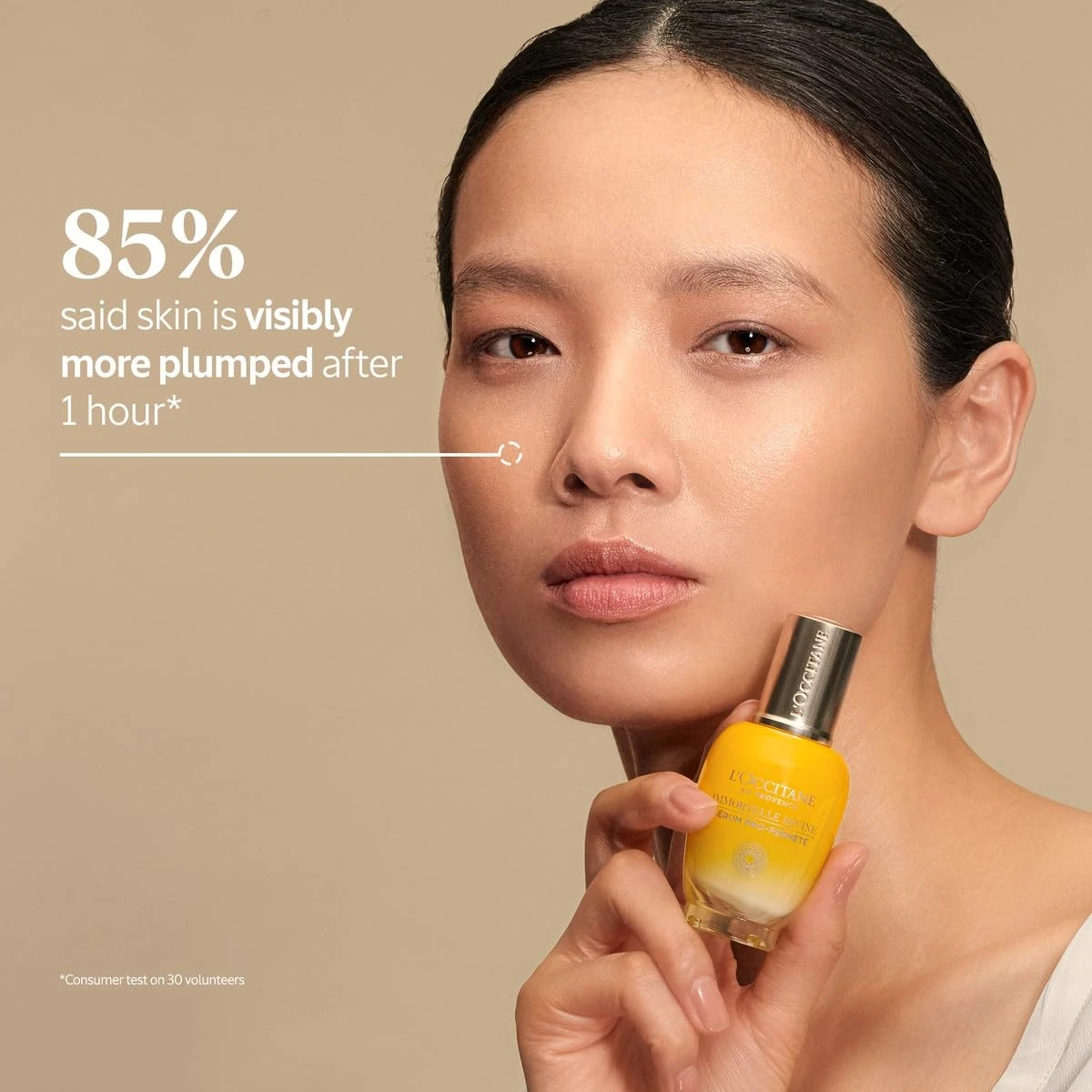 Model holding L'Occitane Immortelle Divine Serum bottle, with a brand claim that 85% said skin looked more plumped after 1 hour in a consumer test of 30 volunteers